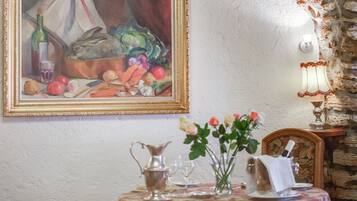 Une table à manger recouverte d’une nappe à motifs floraux, un vase rempli de fleurs et, au mur, un tableau représentant des légumes et une bouteille.