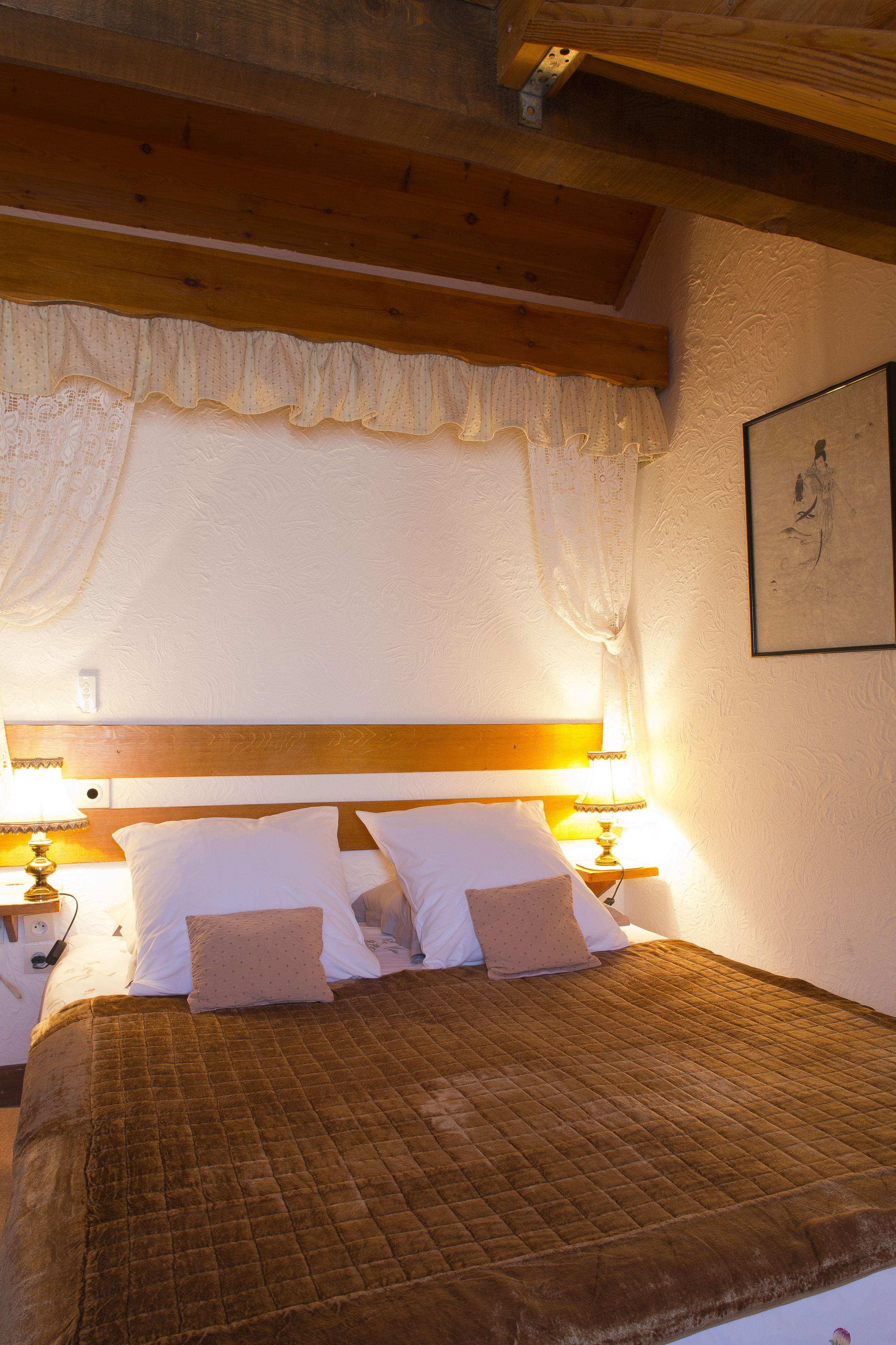 Un lit avec une couette marron, des oreillers blancs et une tête de lit en bois. On y trouve deux lampes de chevet et un tableau encadré accroché au mur.