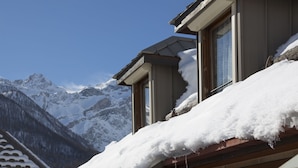 A mountain cabin with a snow-covered roof and windows overlooking a snowy mountain range.