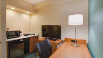 A compact hotel room with a kitchenette, a flat-screen TV, a desk, and a lamp.