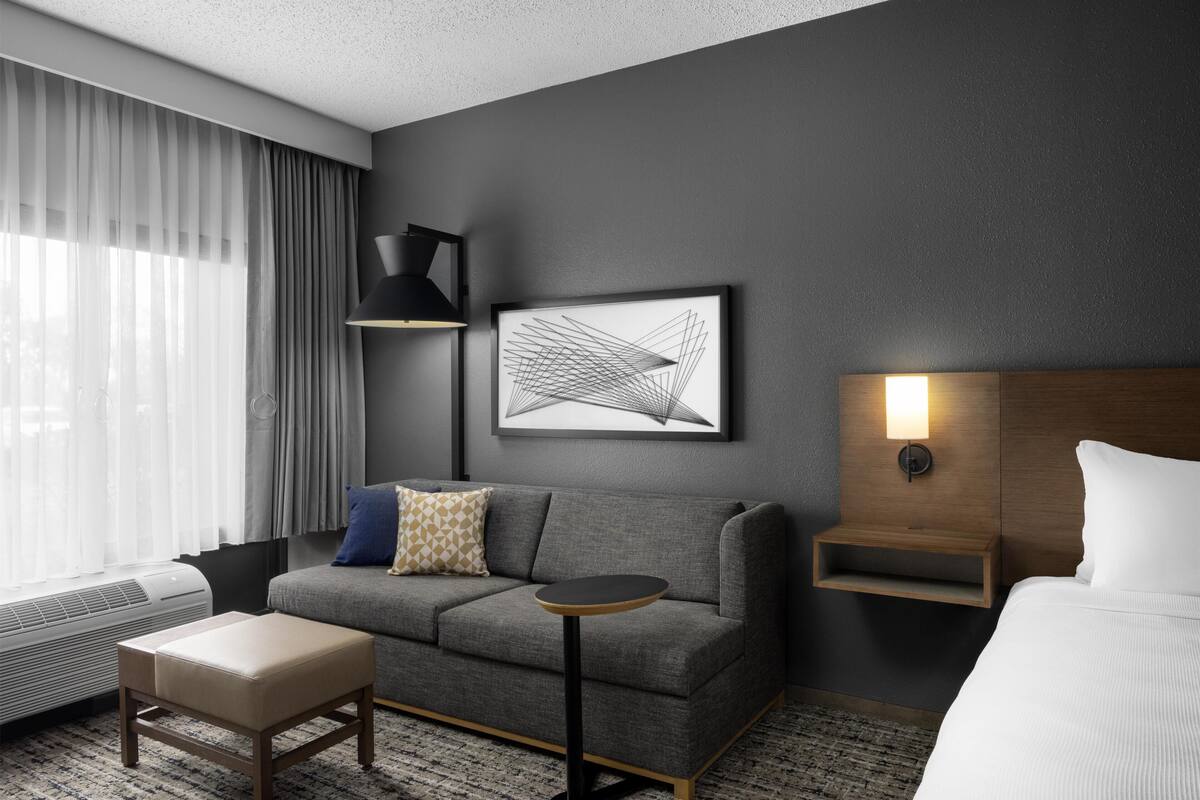 A hotel room with a grey sofa, a small round table, a bedside table with a lamp, a framed abstract artwork, and a bed with white bedding.
