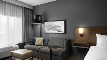 A hotel room with a grey sofa, a small round table, a bedside table with a lamp, a framed abstract artwork, and a bed with white bedding.