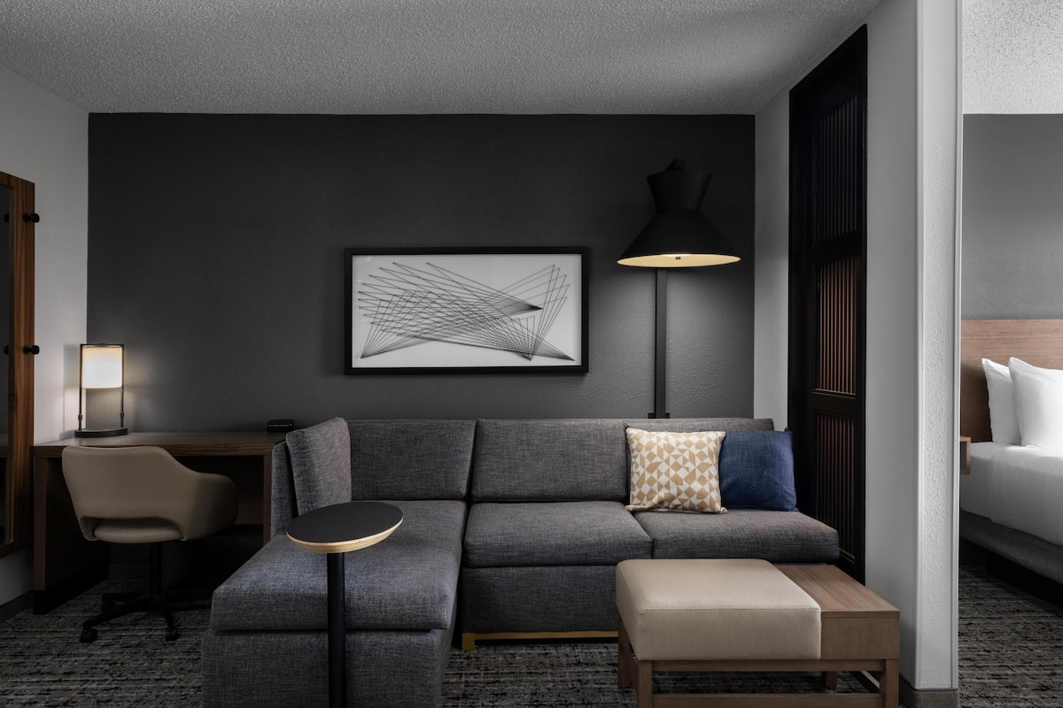 A modern hotel room with a grey sofa, a small round table, a desk with a lamp, and a bed with white bedding.