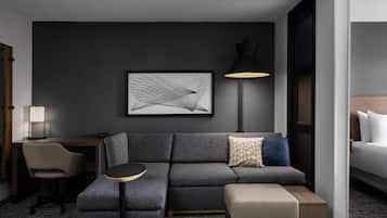 Una habitación de hotel moderna con un sofá gris, una mesita redonda pequeña, un escritorio con lámpara y una cama con ropa de cama blanca.