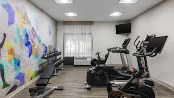 Une salle de sport équipée de matériel d’exercice, d’un banc, d’un téléviseur fixé au mur et d’une fenêtre avec des rideaux.