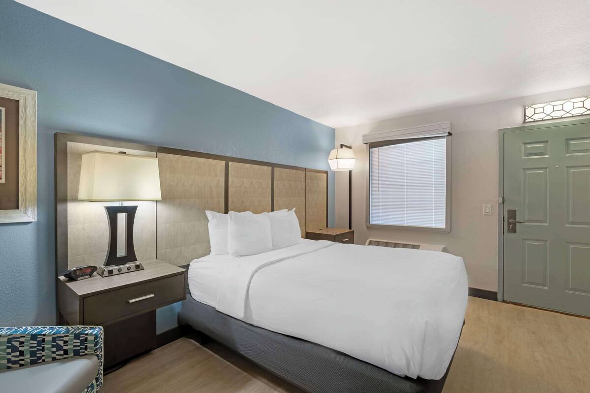 A hotel room with a bed, bedside table, lamp, and a window with blinds.