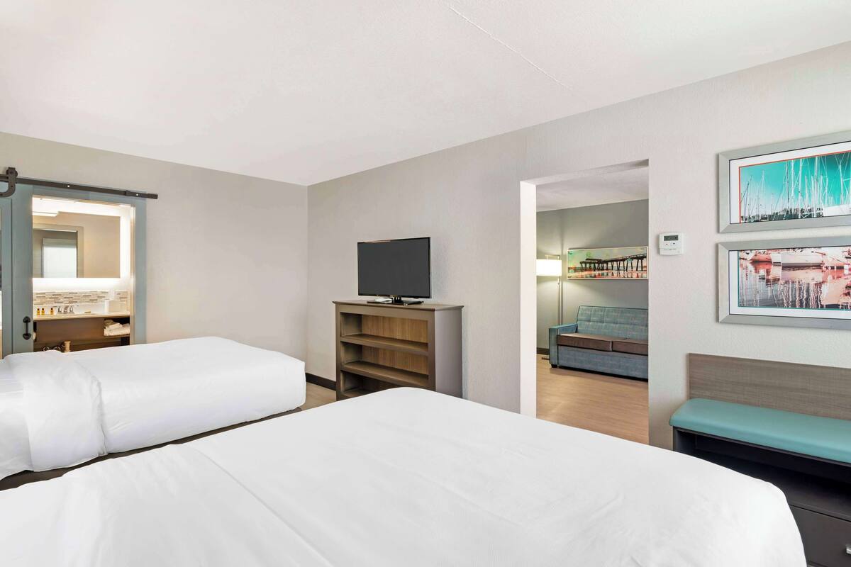 A hotel room with two beds, a television, a desk with a computer, and a bathroom visible through an open door.