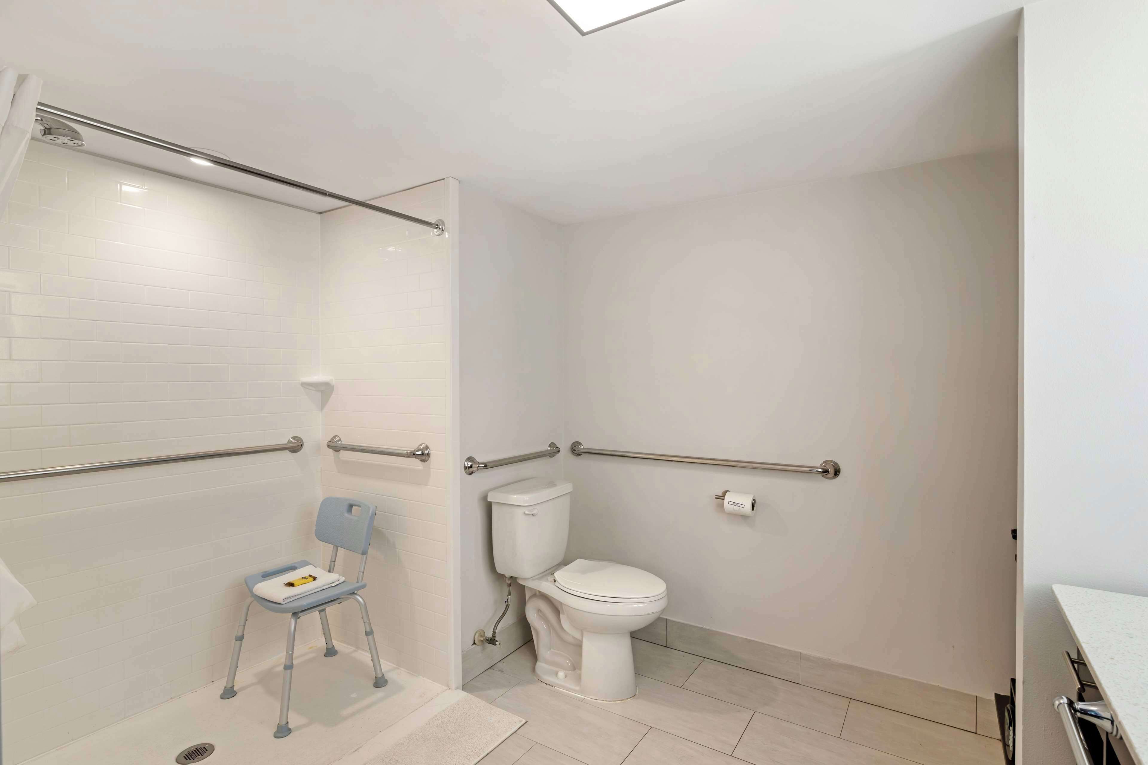 Une salle de bain avec des toilettes blanches, un espace douche équipé d’une barre d’appui et un siège accessible aux personnes en fauteuil roulant.