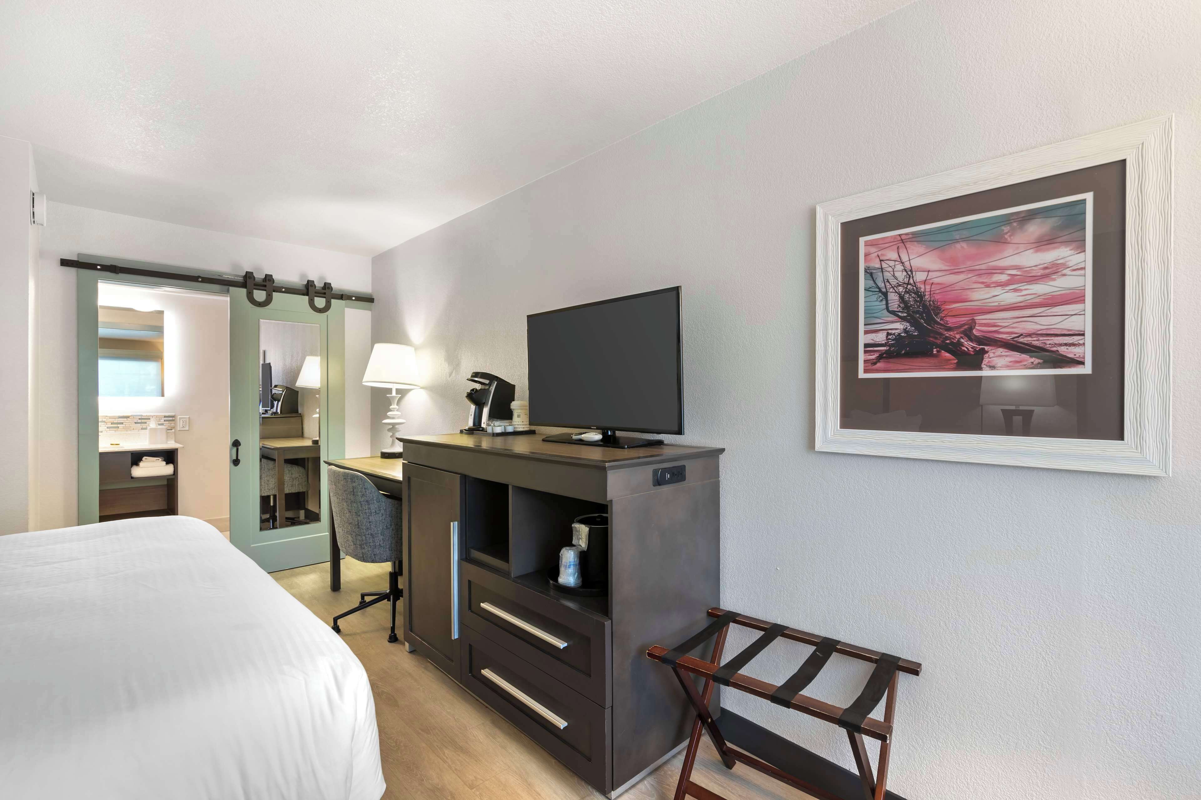 A hotel room with a bed, a desk with a TV, a chair, a lamp, and a framed picture on the wall.