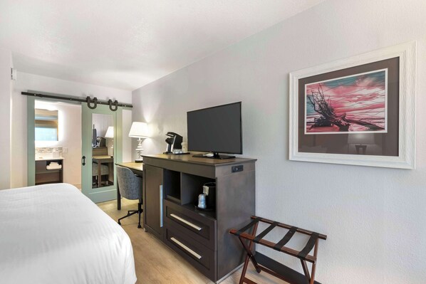 Habitación de hotel con cama, escritorio con televisor, una silla, lámpara y un cuadro en la pared.