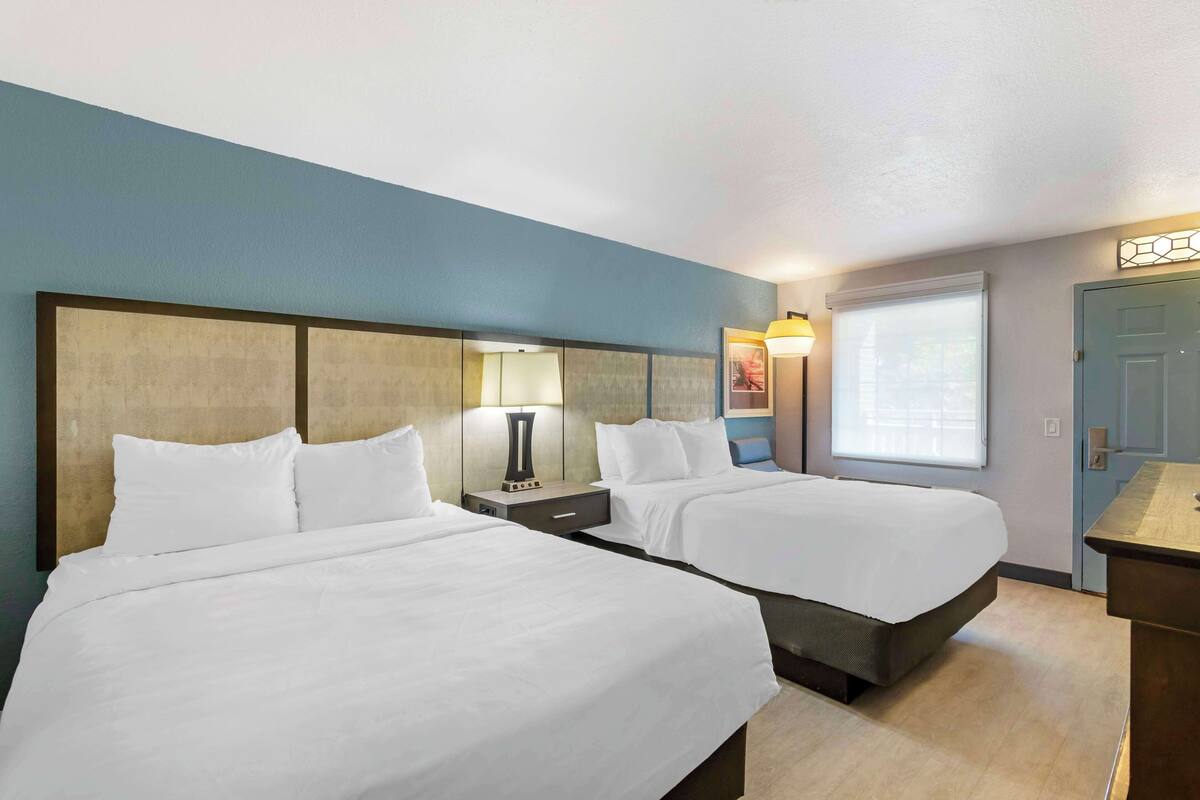 A hotel room with two beds, a nightstand with a lamp, a window with blinds, and a door.