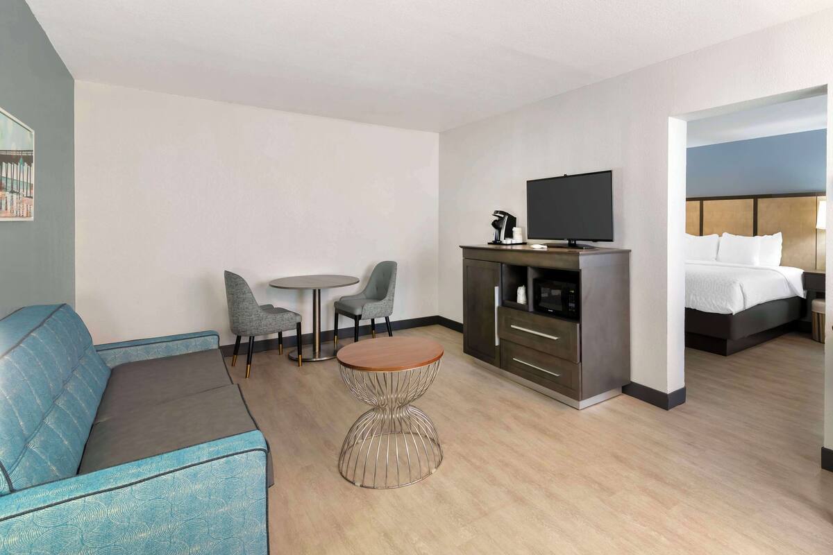 A modern hotel room with a sofa, a small round table, two chairs, a television, a kitchenette, and a bed.