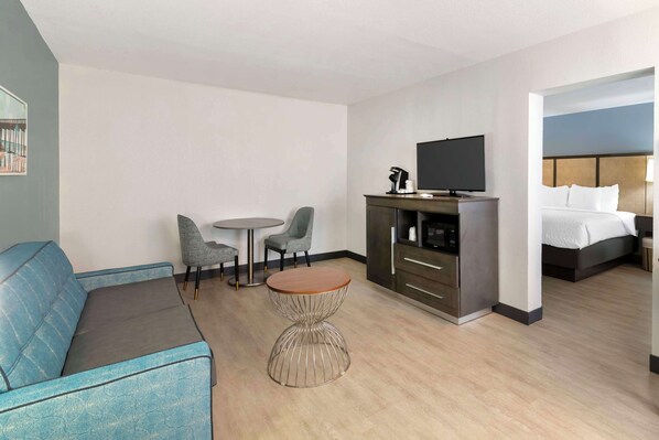 A modern hotel room with a sofa, a small round table, two chairs, a television, a kitchenette, and a bed.