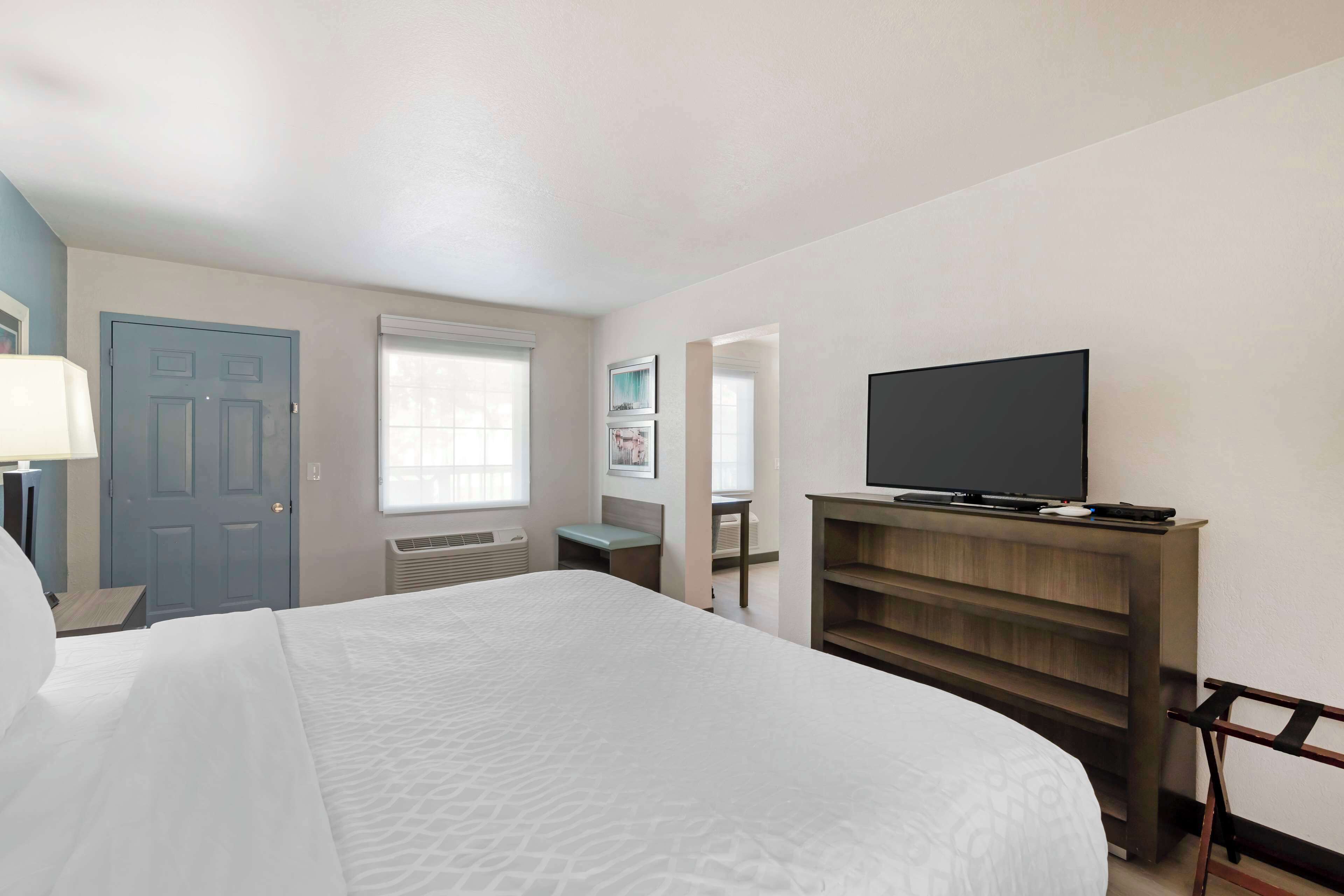 A hotel room with a large bed, a TV on a wooden stand, a desk, and a window with blinds.