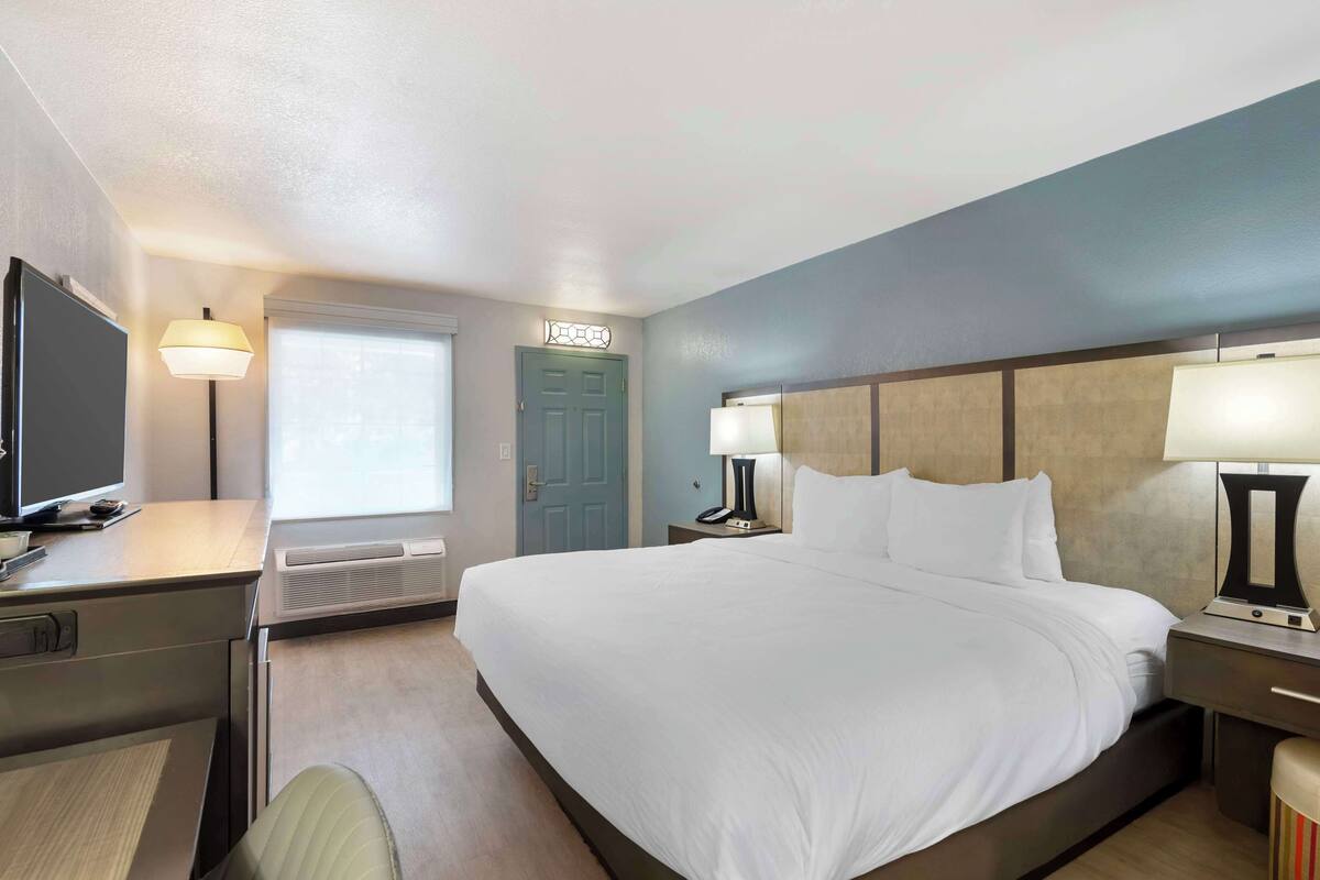 A hotel room with a large bed, bedside tables with lamps, a desk with a computer, a television, and a window with blinds.