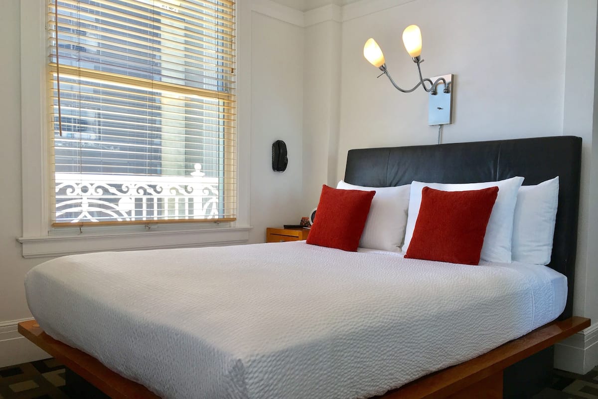 Una cama bien hecha con ropa blanca y almohadas rojas, una mesita de noche con una lámpara y una ventana con persianas.