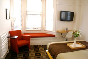 A hotel room with a bed, a red armchair, a small table, a TV mounted on the wall, and a window with blinds.