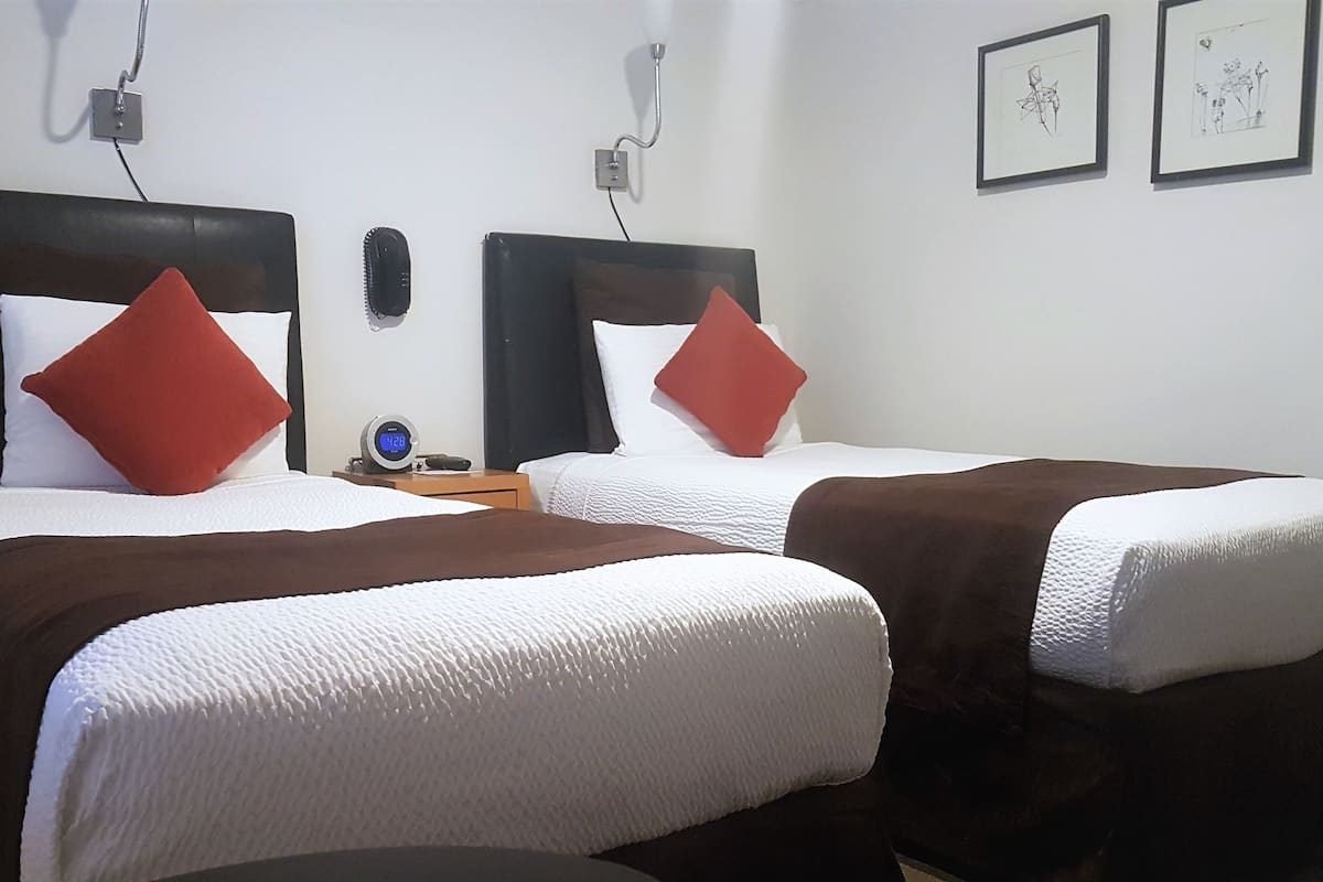 Habitación de hotel con dos camas, cada una con un cojín rojo y una manta marrón. Hay una mesita de noche con un reloj y un teléfono.