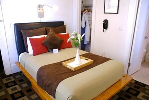 Un lit bien fait, avec une tête de lit marron, une table de chevet ornée d’un vase de fleurs et un téléphone suspendu au mur.