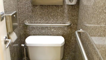 A small, compact bathroom with a toilet, a trash bin, a hand dryer, and a grab bar.