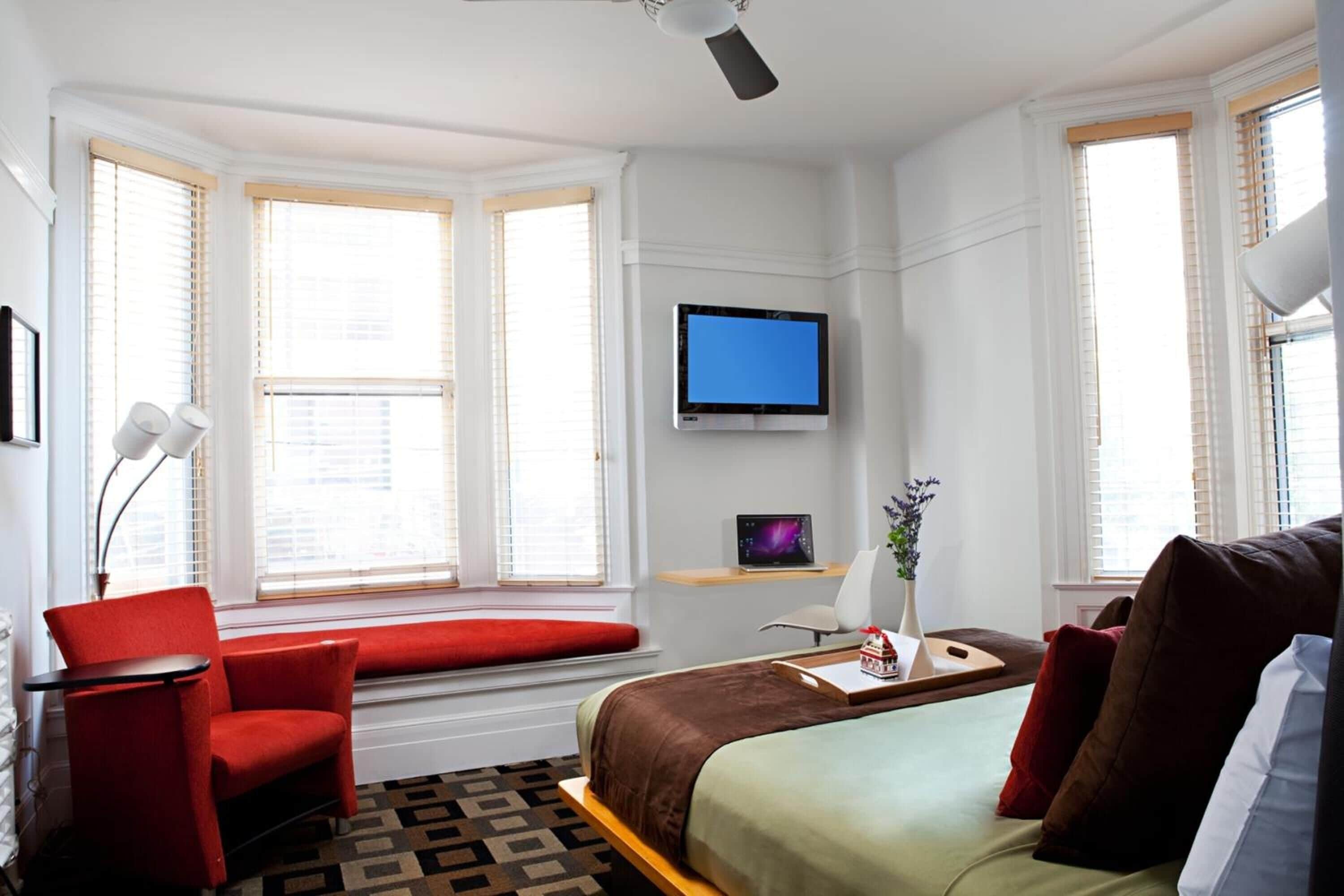 A hotel room with a bed, a red armchair, a desk with a computer, a TV, and large windows with blinds.