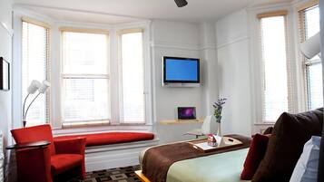 A hotel room with a bed, a red armchair, a desk with a computer, a TV, and large windows with blinds.