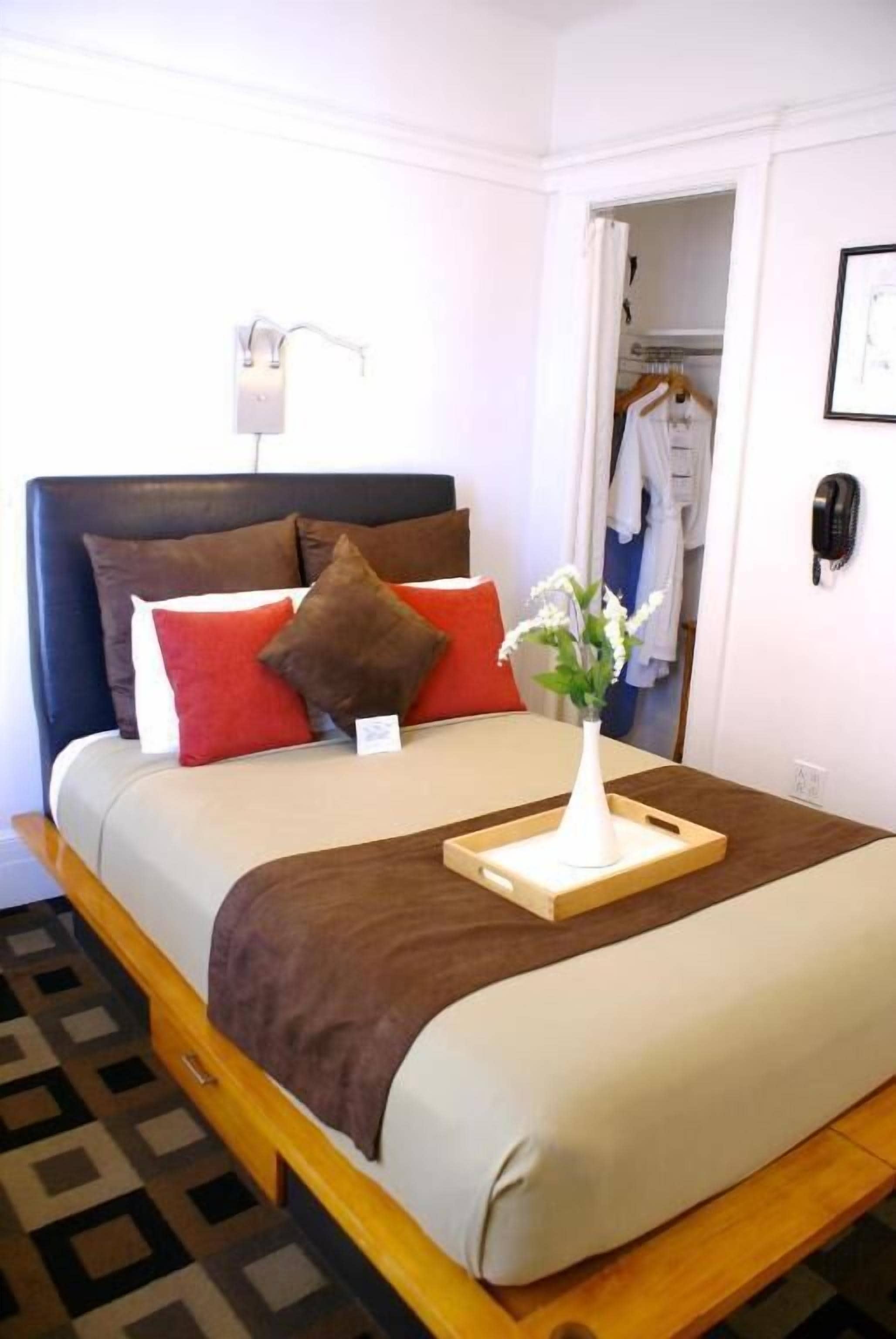A neatly made bed with a brown headboard, a bedside table with a tray holding a vase of flowers, and a closet with hanging clothes.