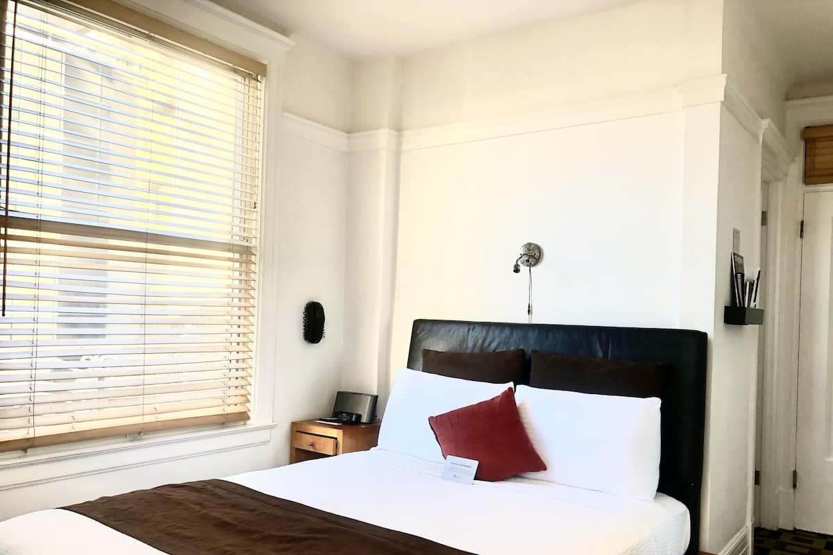 Una habitación de hotel con cama, mesita de noche, lámpara de pared y ventana con persianas.