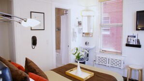 A hotel room with a bed, a bedside table with a tray, a lamp, a bathroom with a sink, and a window with blinds.