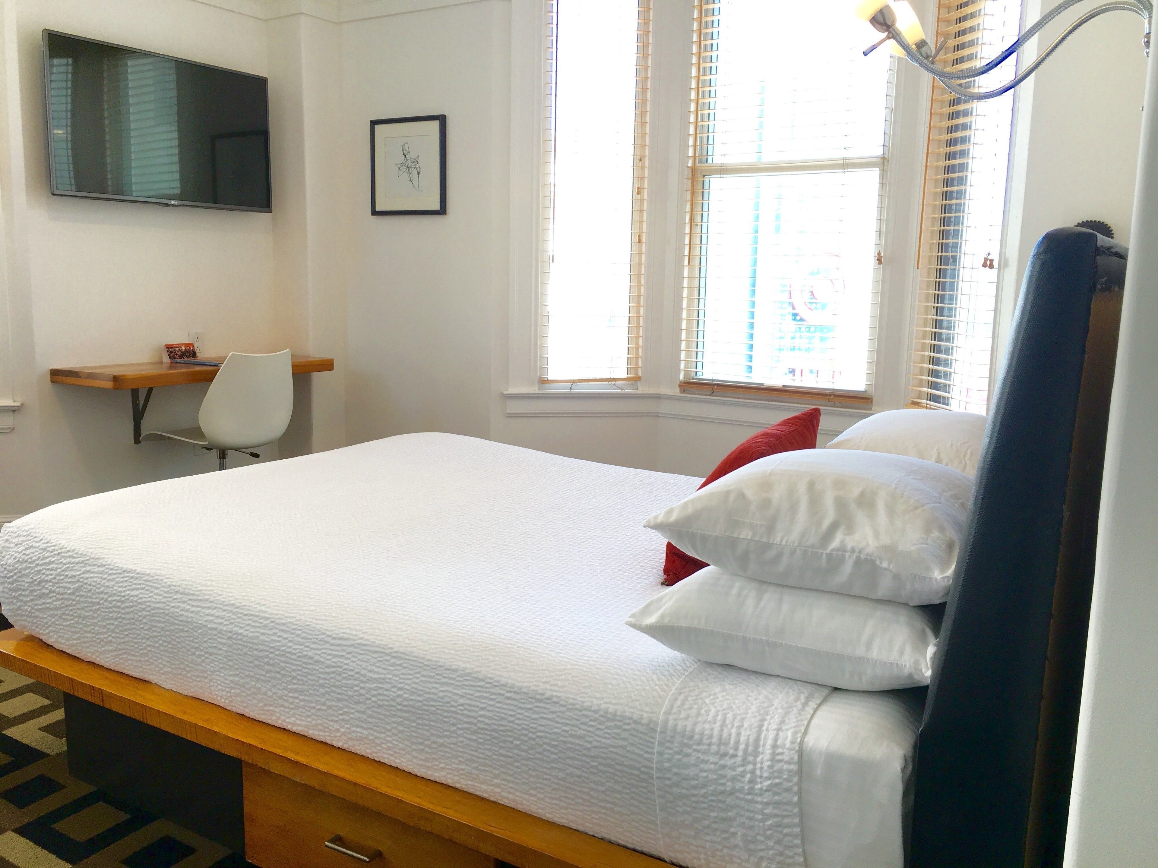 A bedroom with a bed, a desk, a chair, a window with blinds, and a framed picture on the wall.
