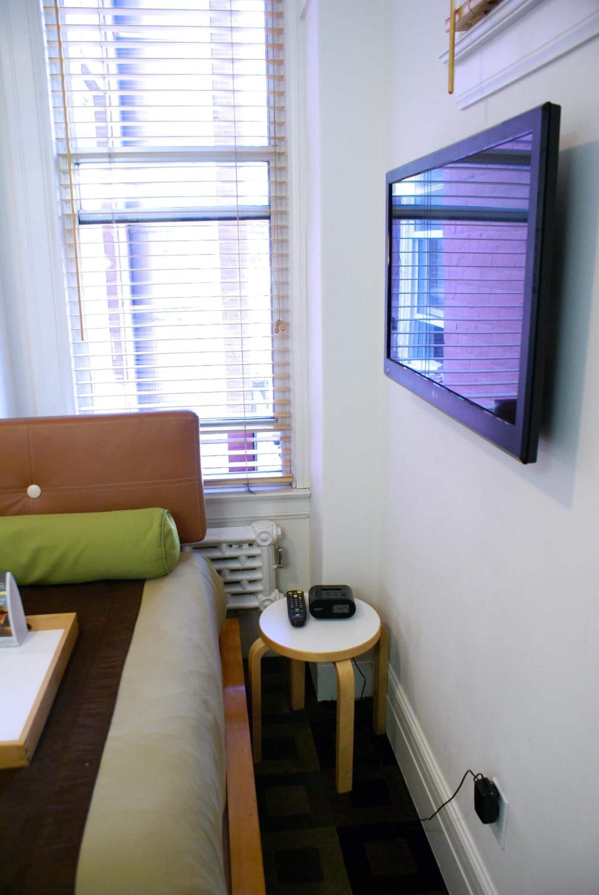 A small room with a bed, a small table, a wall-mounted TV, and a window with blinds.
