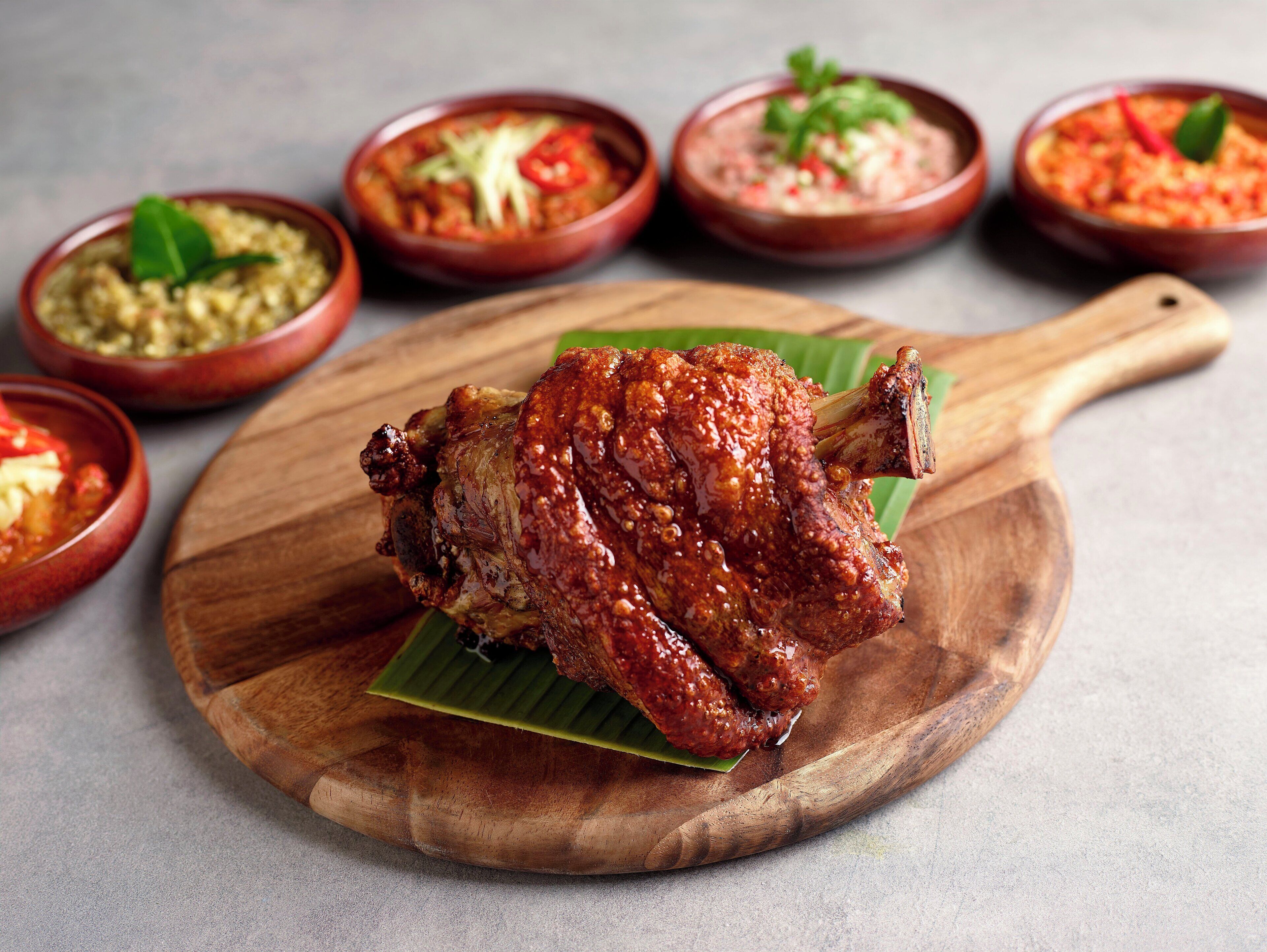 A roasted chicken on a wooden board with a banana leaf, surrounded by small bowls of sauces.