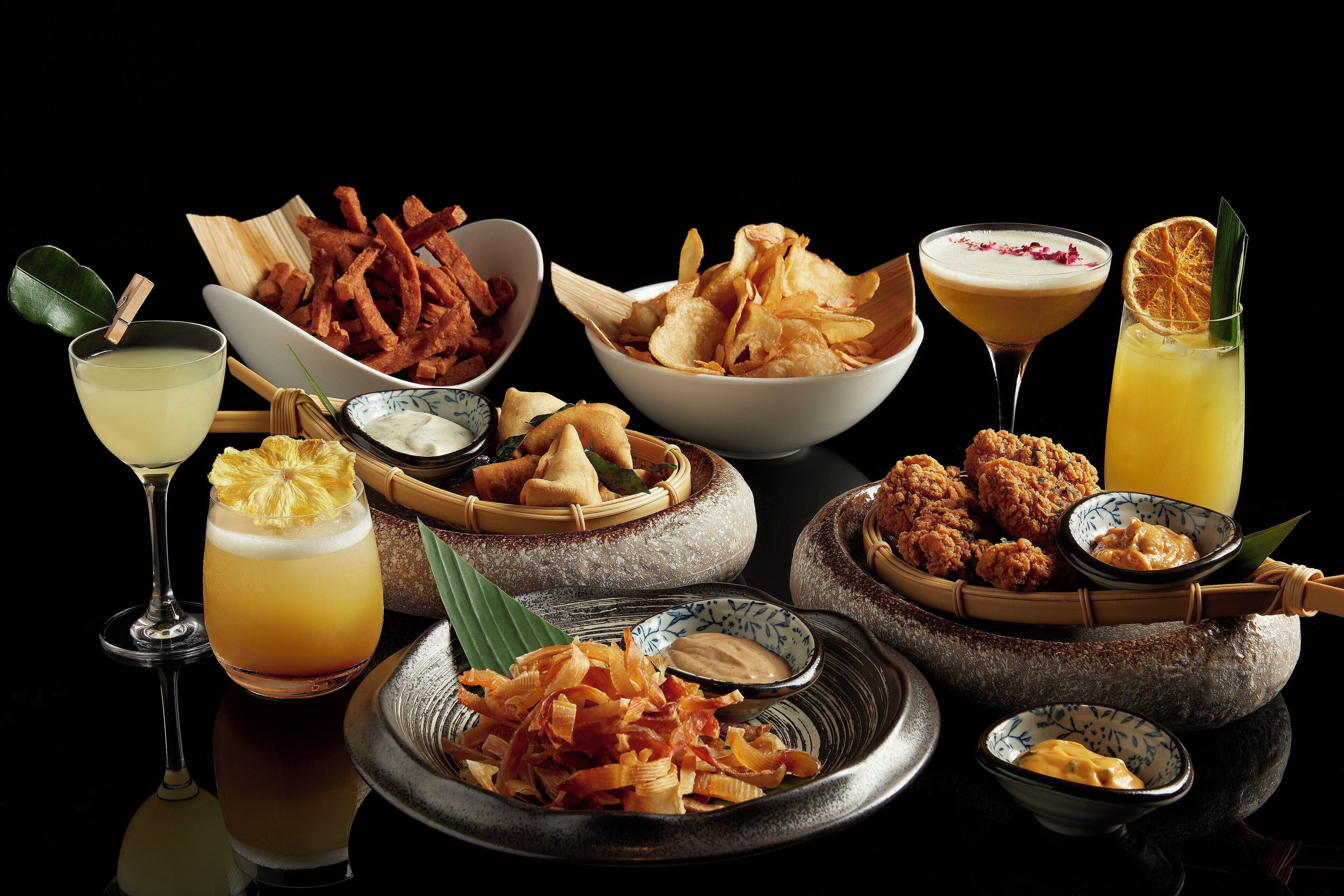 A variety of food and drinks, including fried snacks, a cocktail, and a fruit drink, arranged on a reflective surface.