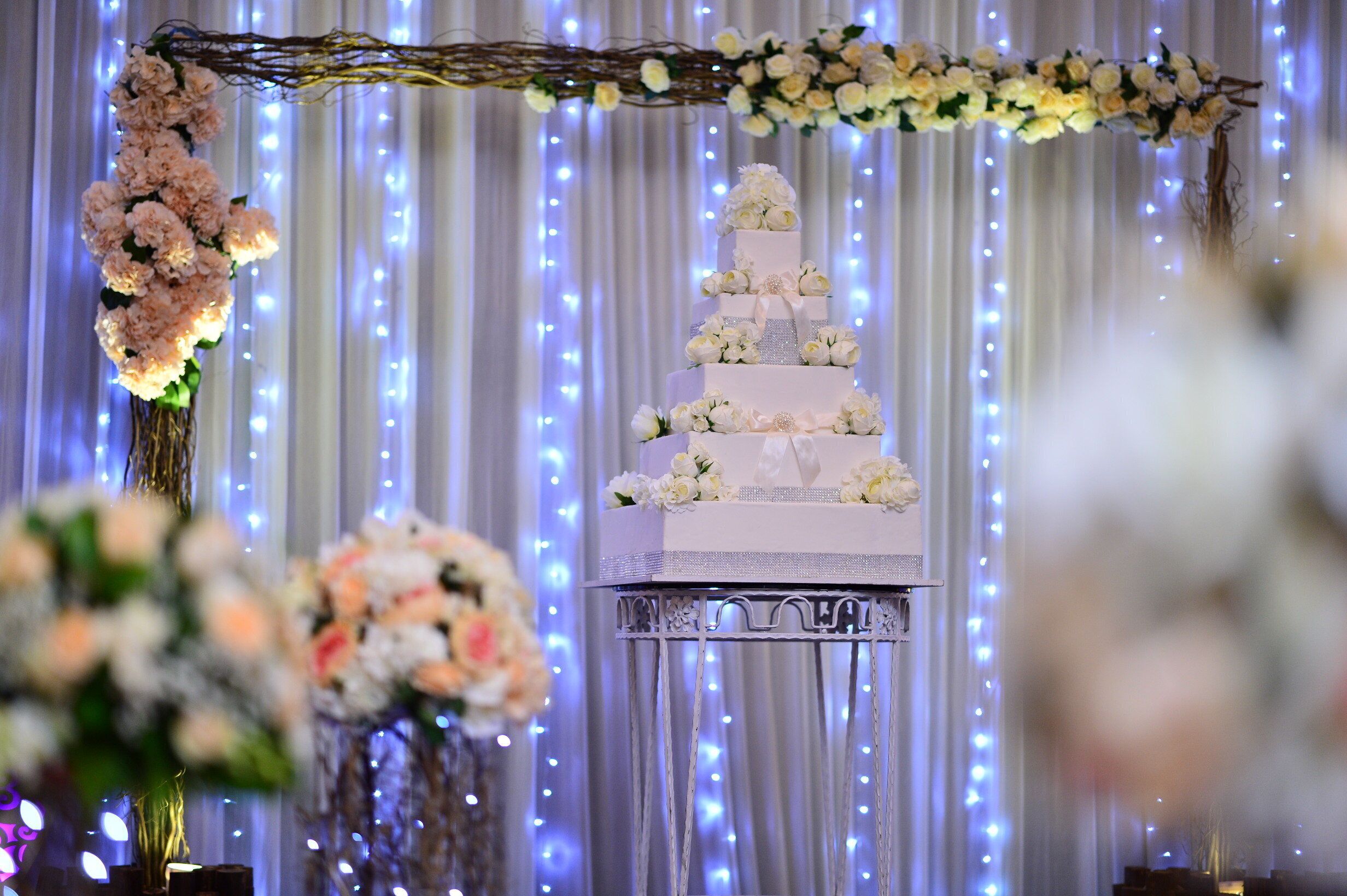 Un gâteau de mariage à plusieurs étages, posé sur un présentoir, agrémenté de compositions florales et illuminé de guirlandes lumineuses en arrière-plan.
