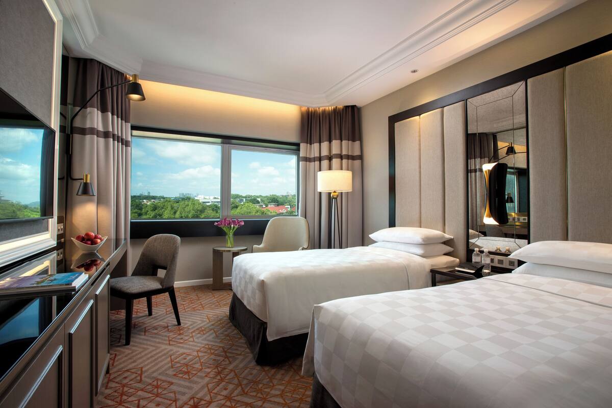 Ein Hotelzimmer mit zwei Betten, einem Schreibtisch mit Computer, einem Stuhl, einem kleinen Tisch mit Blumenvase und einem Fenster mit Blick auf die Stadt.