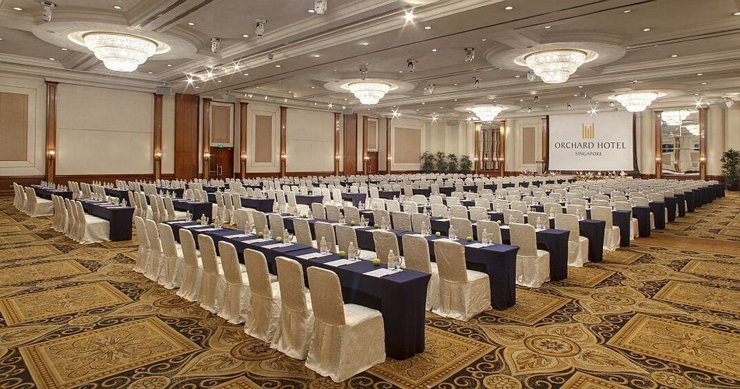 Un salón de conferencias en el Orchard Hotel, Singapur, con filas de mesas y sillas, suelo alfombrado y candelabros.