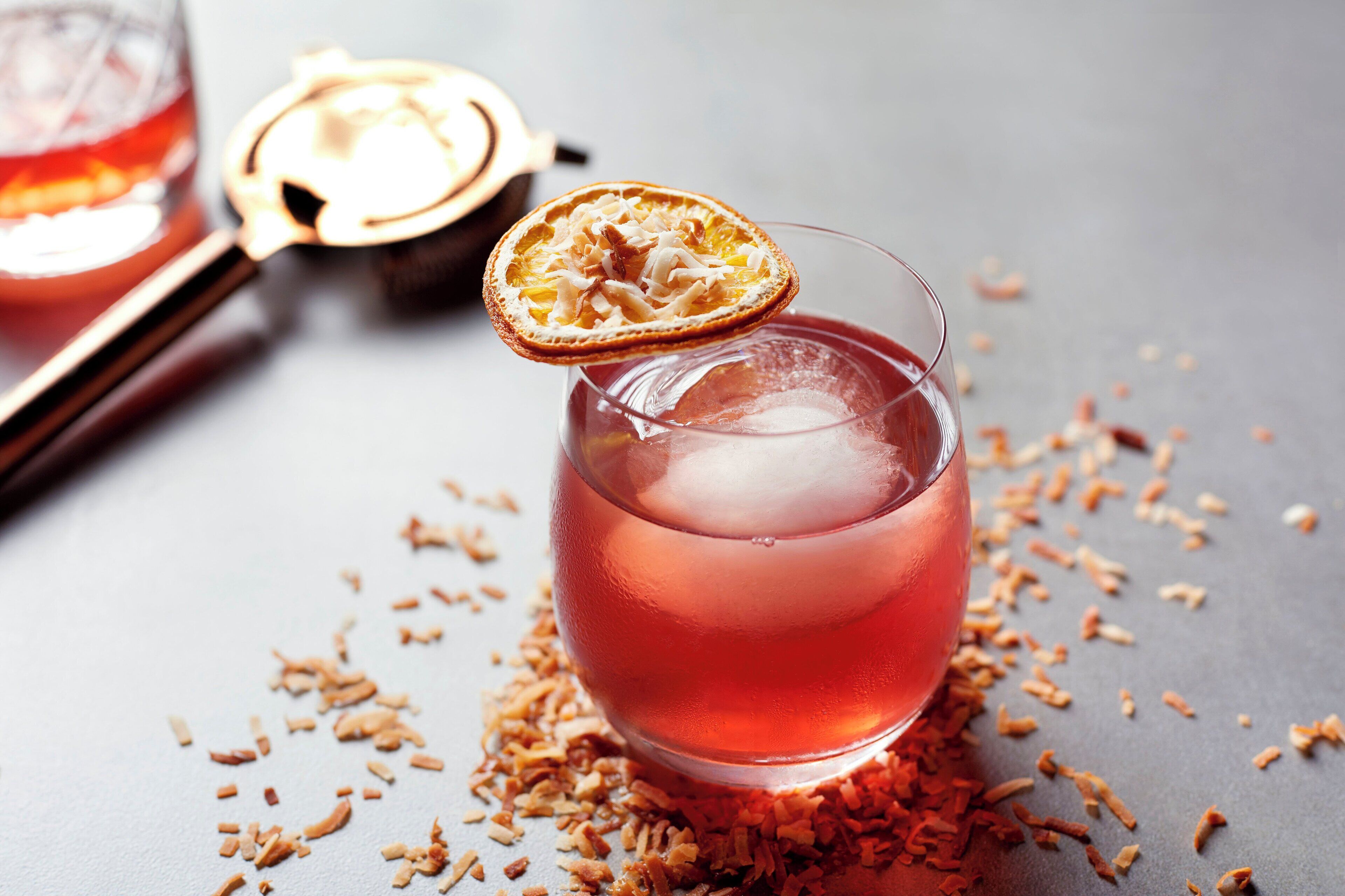 Un vaso de bebida roja con adorno de fruta seca, rodeado de trozos de fruta seca dispersos sobre una superficie gris.