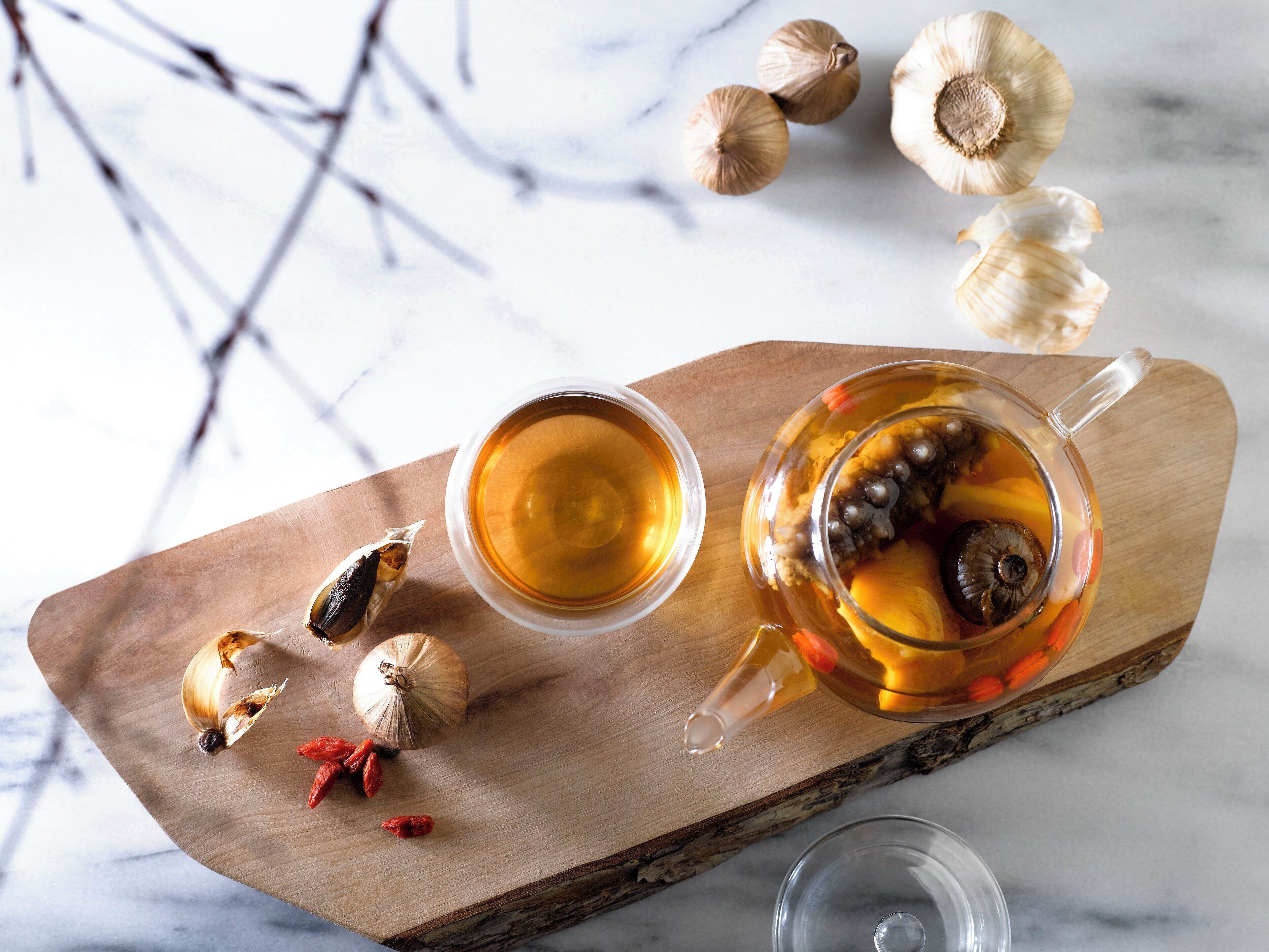A wooden tray with a glass teapot and cup, garlic bulbs, and red goji berries.
