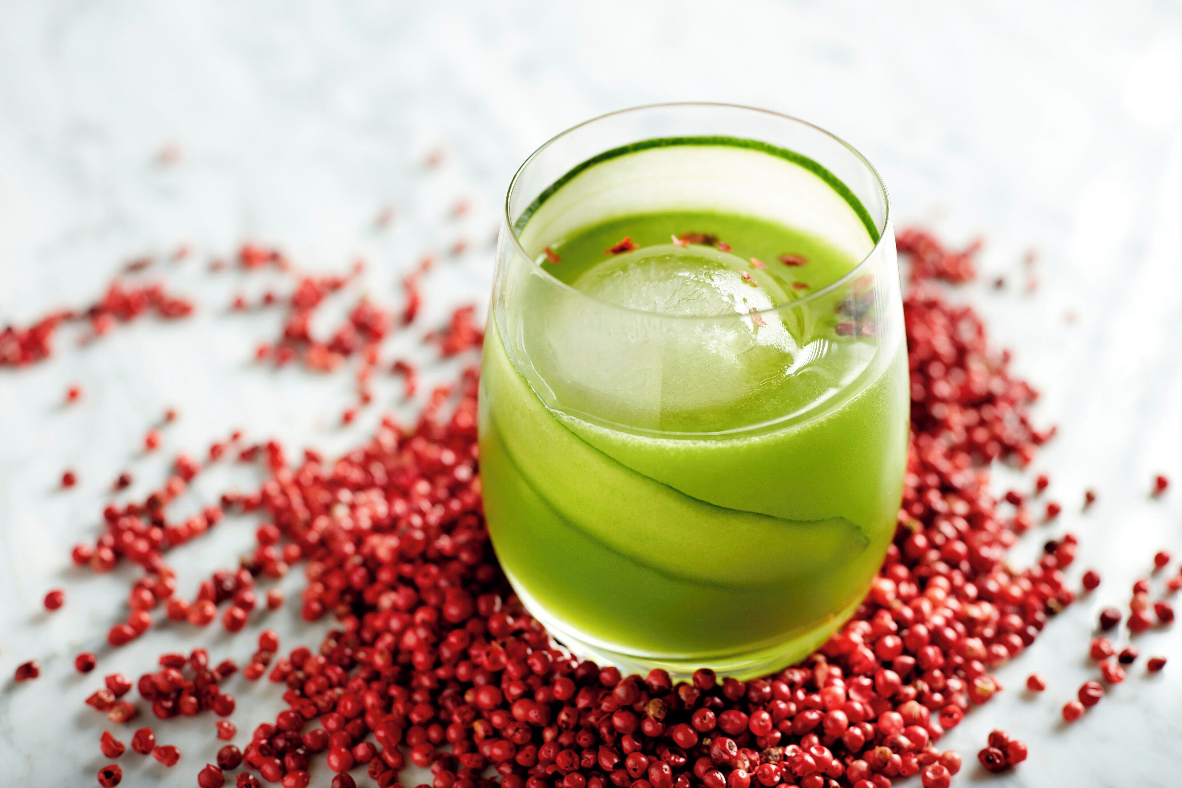 A glass of green beverage surrounded by red berries on a white surface.