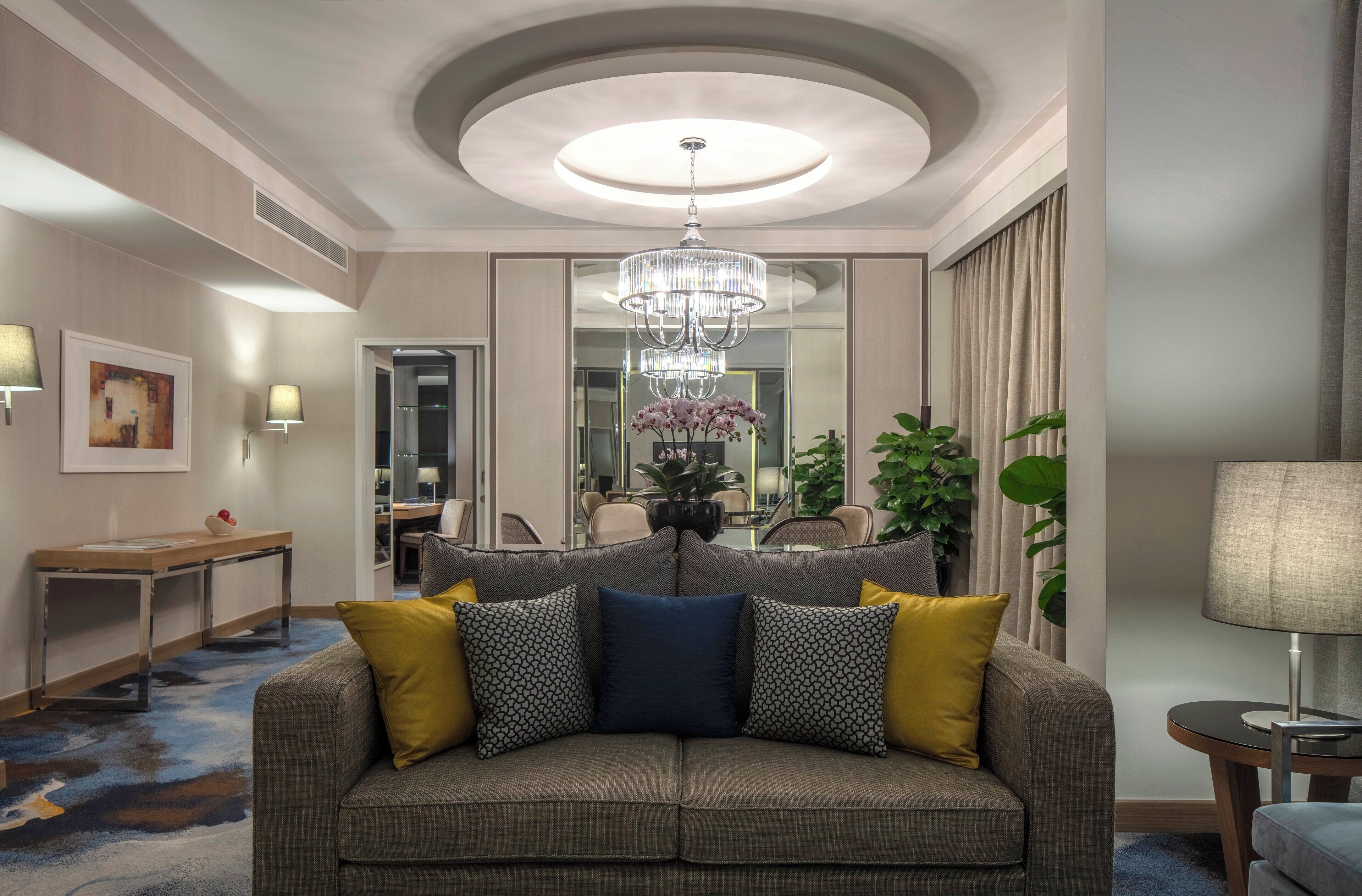 A modern living room with a grey sofa, patterned cushions, a chandelier, and a large mirror reflecting the room.