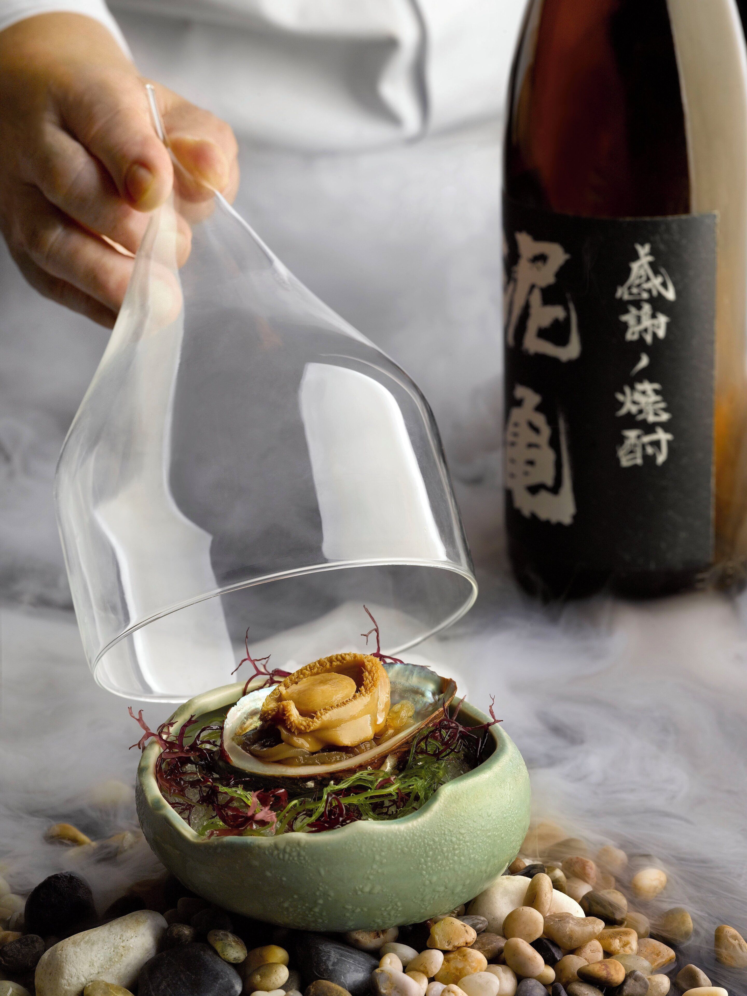 A person holding a clear plastic dome over a dish with food and a bottle in the background.