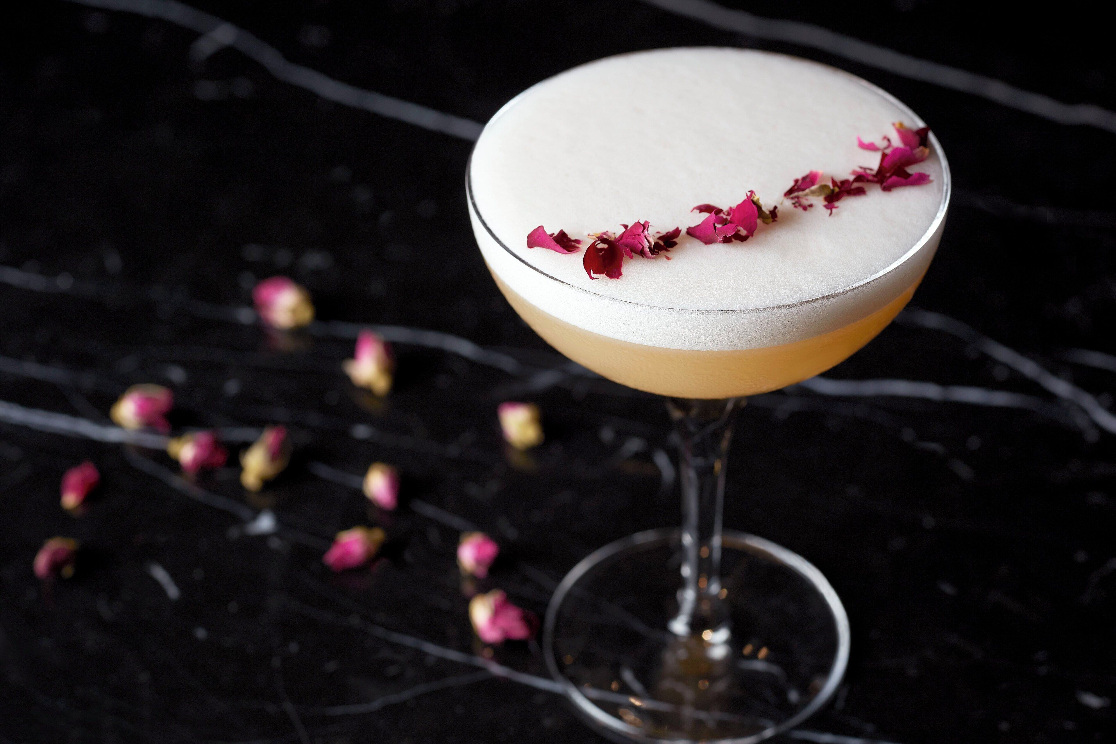 A cocktail in a stemmed glass with a layered appearance and a garnish of pink petals.