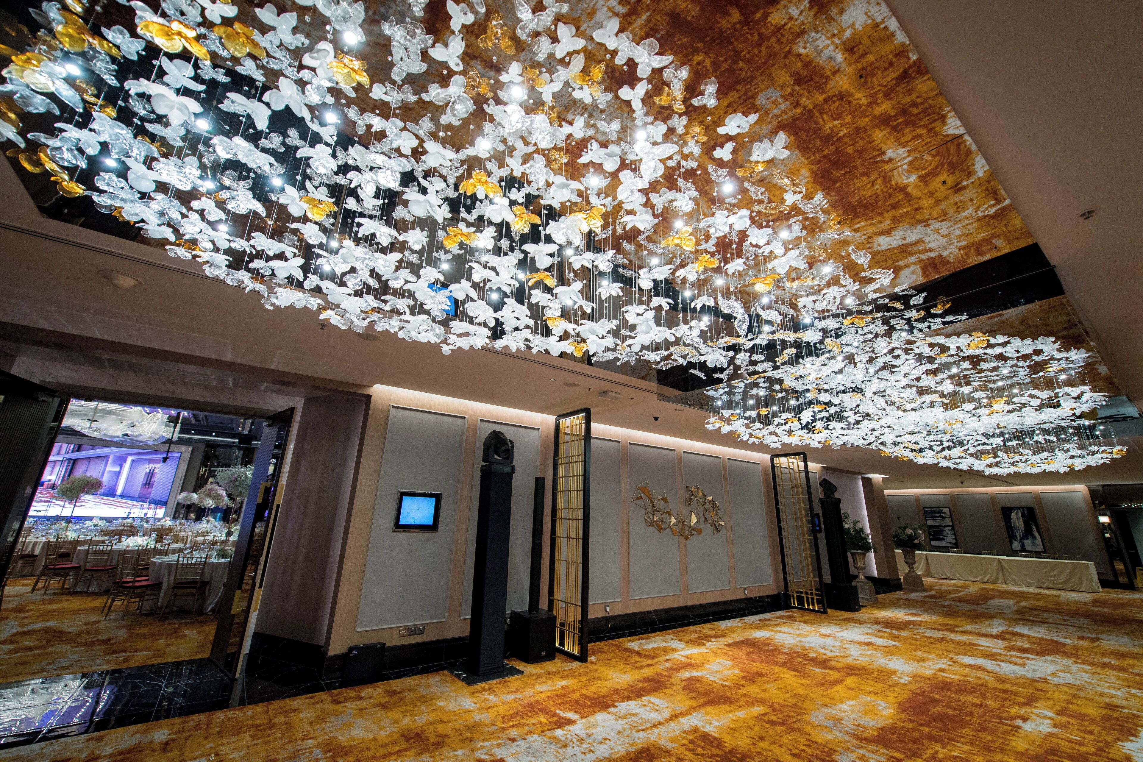A spacious lobby with a high ceiling decorated with hanging floral arrangements, a reception area with a digital screen, and a seating area with tables and chairs.