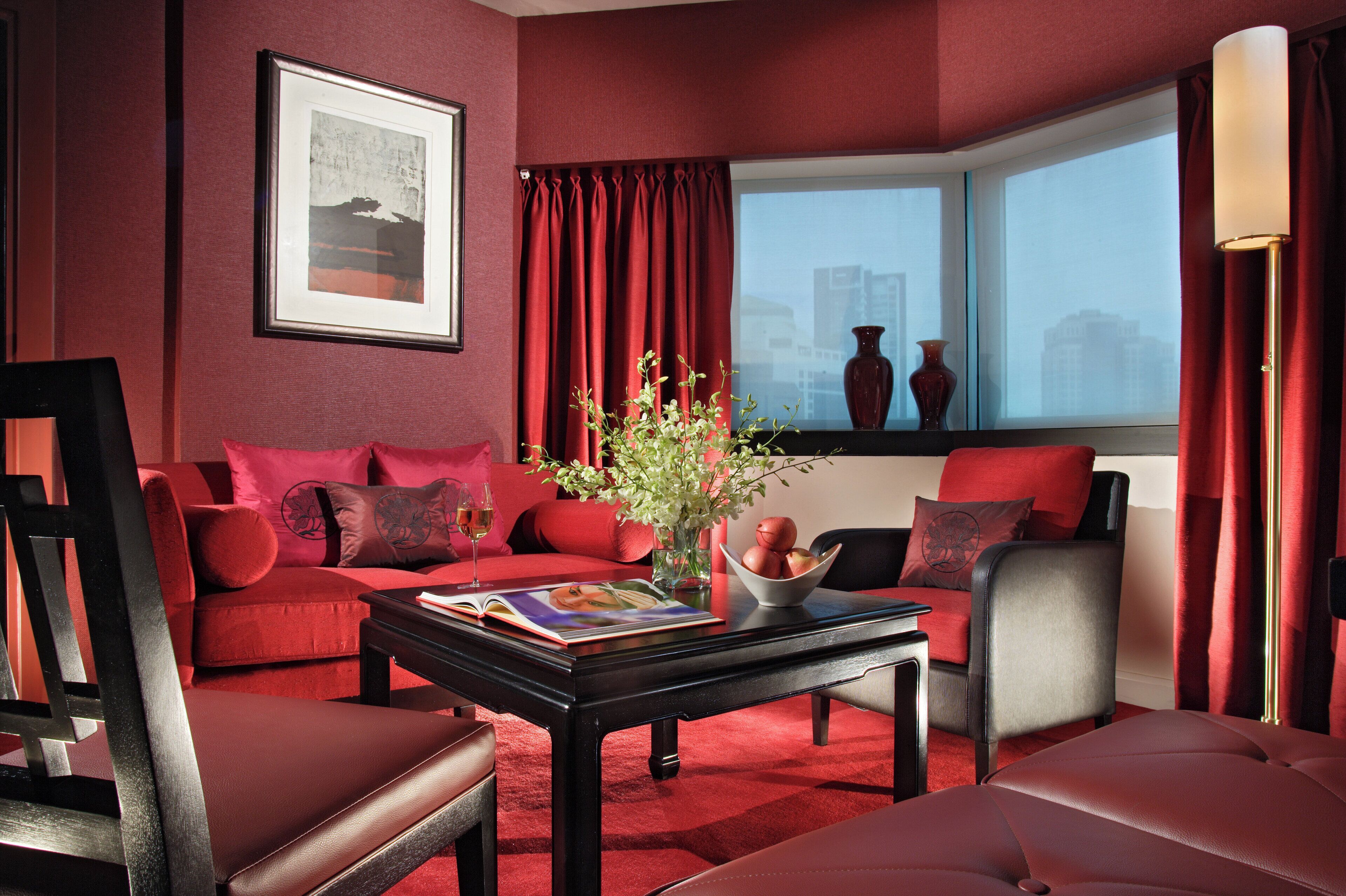 A modern living room with red walls, a sofa, a coffee table with a magazine, a vase with flowers, and a window with curtains.