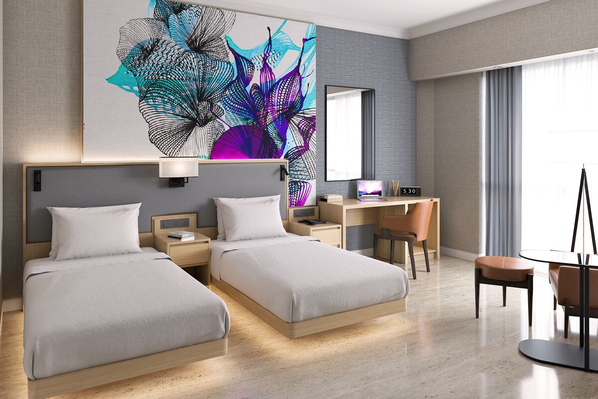 A modern hotel room with two beds, a desk, a chair, a laptop, and a large abstract painting on the wall.