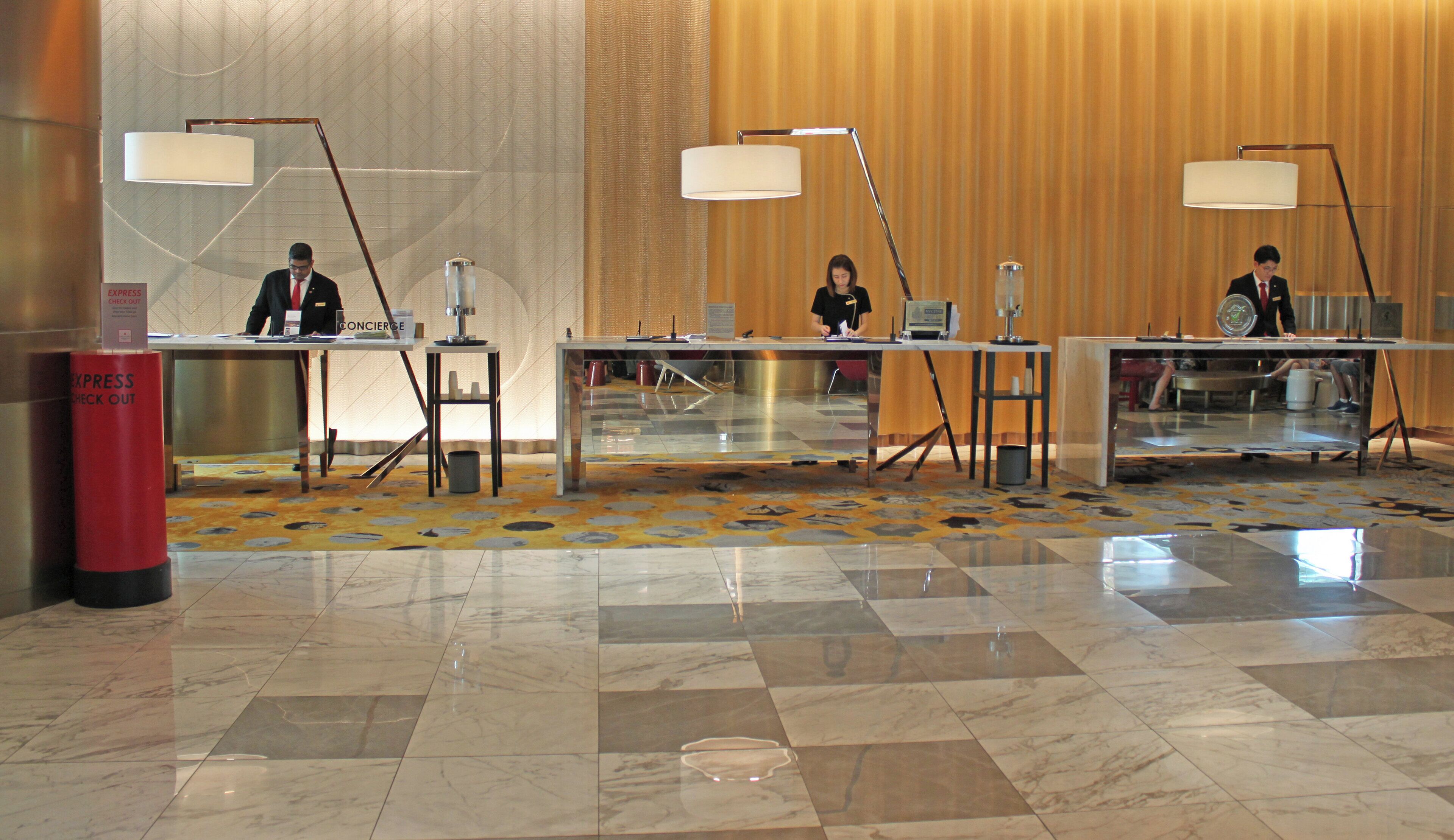 Zona de recepción de un hotel con tres mostradores, cada uno equipado con una computadora y un teléfono. Dos lámparas de pie grandes se encuentran encima de los mostradores, y se ve un basurero rojo a la izquierda. El piso está cubierto con un patrón geométrico.