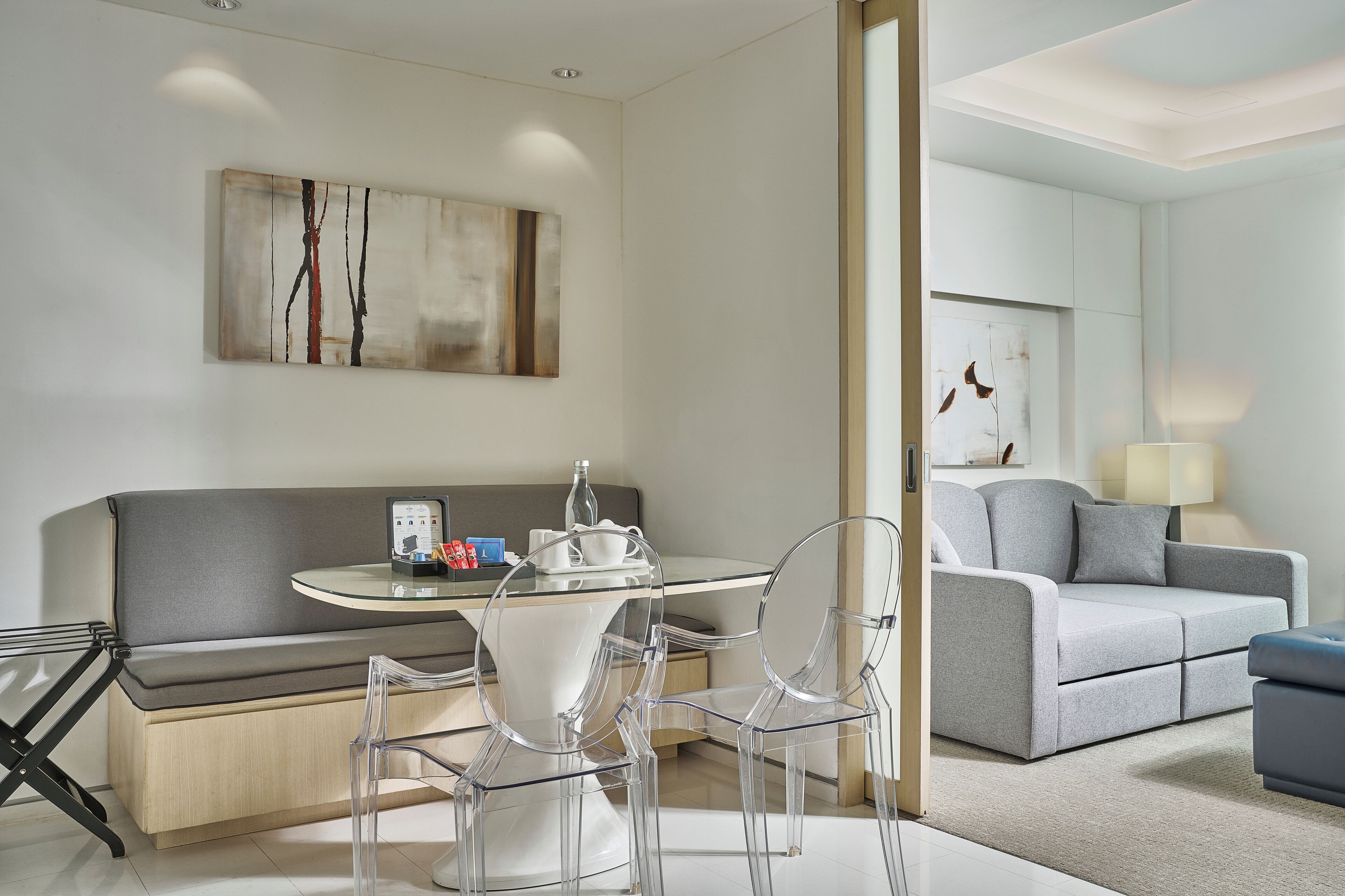 Ein modernes Wohnzimmer mit einem grauen Sofa, einem Glastisch und transparenten Stühlen. Ein Gemälde hängt an der Wand, und auf einem Beistelltisch steht eine Lampe.