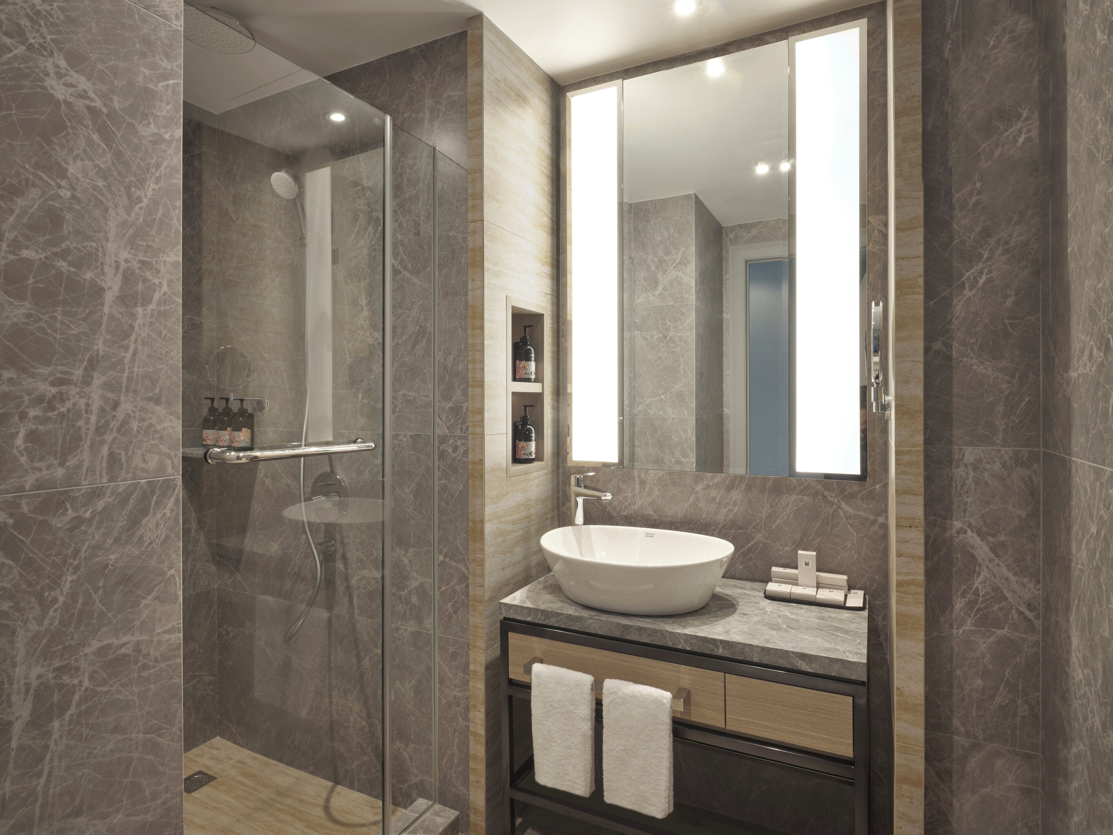 Un baño moderno con ducha envuelta en vidrio, un espejo grande y un tocador con loseta de mármol y un lavamanos.