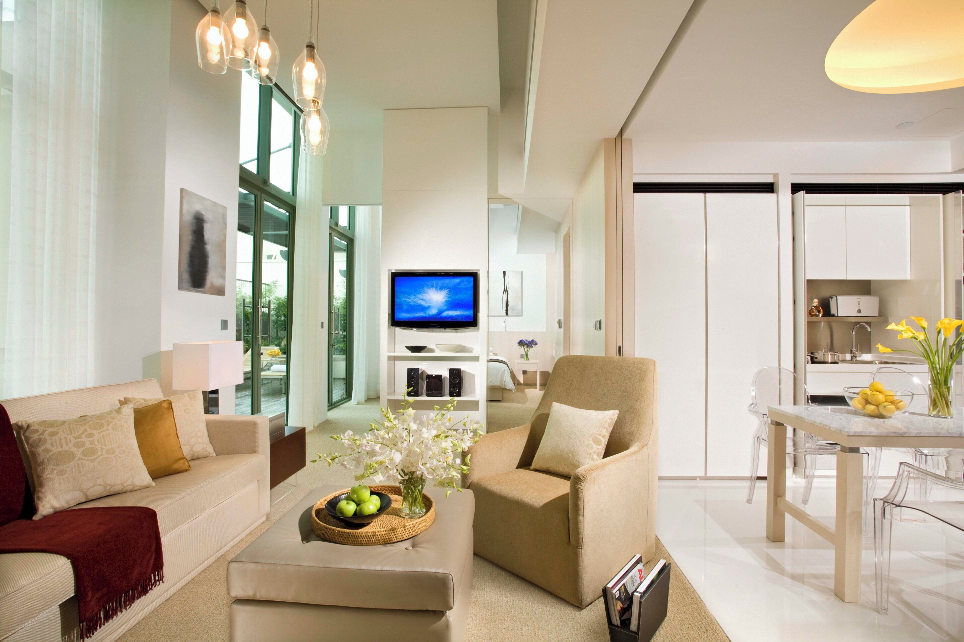 A modern living room with a sofa, armchair, coffee table with fruit and flowers, a TV, and a dining area with a table and chairs.