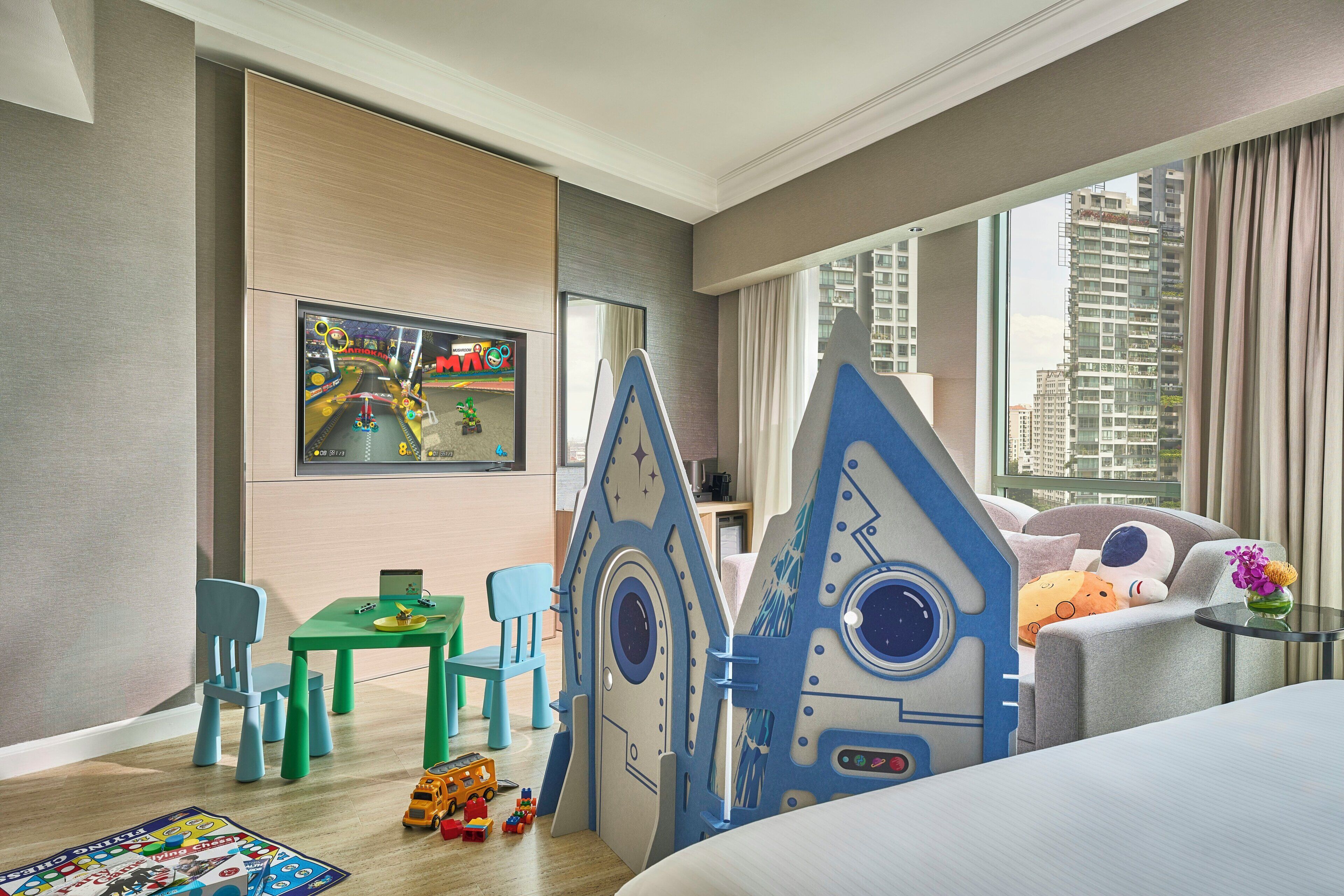 Una habitación de hotel con cama, un área de juegos con una estructura espacial y una mesa verde, además de una ventana con vista a la ciudad.